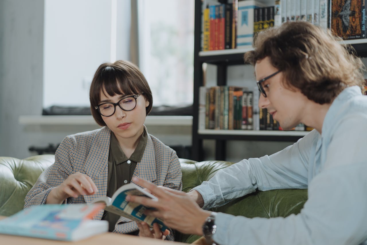 pexels-cottonbro-4861395 Eine Frau und ein Mann sitzen nebeneinander auf einem Sofa und lesen gemeinsam in einem Buch.